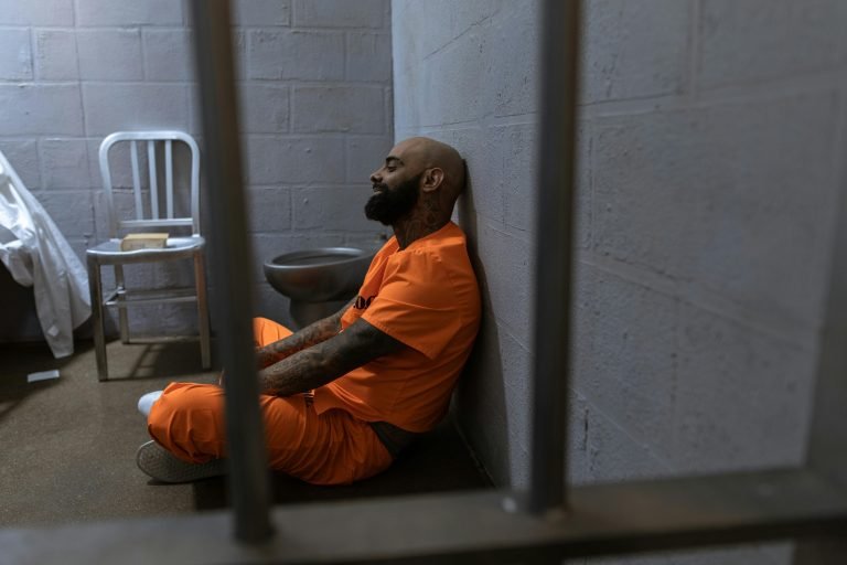 Home A bald man in an orange jumpsuit sits on the floor of a jail cell, leaning against a concrete wall.